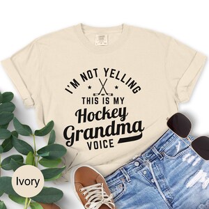 May include: Ivory-colored t-shirt with the text "I'M NOT YELLING THIS IS MY Hockey Grandma VOICE" in black. The design includes crossed hockey sticks and stars. The shirt is paired with denim shorts, sunglasses, and brown shoes.