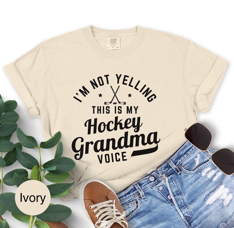 May include: Ivory-colored t-shirt with the text "I'M NOT YELLING THIS IS MY Hockey Grandma VOICE" in black. The design includes crossed hockey sticks and stars. The shirt is paired with denim shorts, sunglasses, and brown shoes.