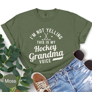 May include: An olive green t-shirt with the text "I'M NOT YELLING THIS IS MY Hockey Grandma VOICE" in white. The shirt features crossed hockey sticks and stars. The shirt is paired with denim shorts, brown shoes, and sunglasses.