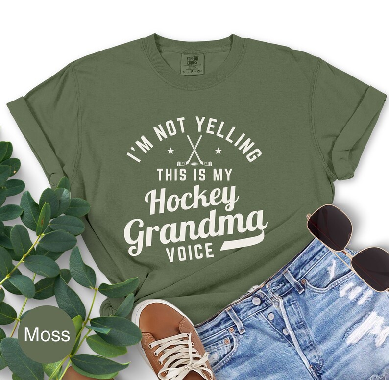 May include: An olive green t-shirt with the text "I'M NOT YELLING THIS IS MY Hockey Grandma VOICE" in white. The shirt features crossed hockey sticks and stars. The shirt is paired with denim shorts, brown shoes, and sunglasses.