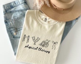 May include: An ivory-colored t-shirt with a black line drawing of physical therapy equipment, including a walker, crutches, and a wheelchair. The words "physical therapy" are printed below the drawings. The shirt is paired with light blue jeans and a tan hat.