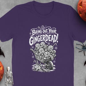 May include: A purple short-sleeve t-shirt featuring a white graphic print of a wheelbarrow filled with gingerbread cookies and the text "Bring out your GINGERDEAD!" surrounded by candy canes and snowflake accents.