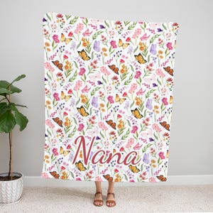 May include: A white blanket with a floral and butterfly pattern. The design features orange, pink, and purple flowers with orange and pink butterflies. The word "Nana" is written in a cursive font across the bottom.