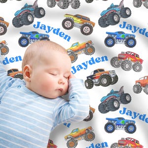 May include: A white fabric blanket featuring a repeating pattern of colorful monster trucks in shades of blue, orange, red, and yellow, interspersed with the name Jayden printed in a bold blue sans-serif font across the surface.