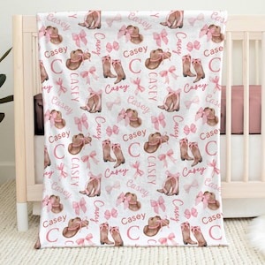 a personalized western themed baby girl blanket draped from a tan crib. The name "Casey" is all throughout the repeating pattern of cowgirl boots with pink bows and cowgirl hats with pink bows.