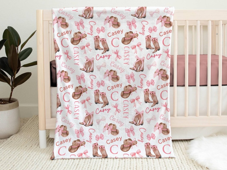 a personalized western themed baby girl blanket draped from a tan crib. The name "Casey" is all throughout the repeating pattern of cowgirl boots with pink bows and cowgirl hats with pink bows.