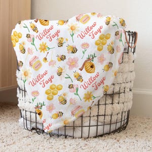 May include: White baby blanket with a repeating pattern of bees, honeycombs, flowers, and the name "Willow Faye" in pink lettering.