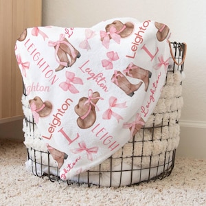 a girly pink bows western blanket is draped on a metal wire basket in a bedroom setting. The design also includes soft, brown cowgirl boots and hat decorated with pink bows.