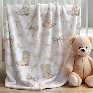 May include: A white fleece blanket featuring a repeating pattern of light brown cowboy boots and decorative bows. The soft-textured fabric displays these illustrations in a consistent, spaced-out arrangement across the entire surface of the blanket.