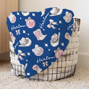 May include: A blue baby blanket with a repeating pattern of cowboy hats, boots, bows, and ornaments. The name "Harlow" is written in white script throughout the pattern.