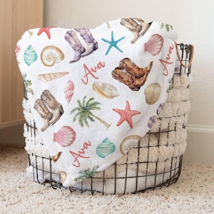 Custom name coastal cowgirl beach pattern blanket. Blanket features jeweled cowgirl boots, beach shells in peaches, turquoise, and cream colors, and personalized girl's name in a salmon color.