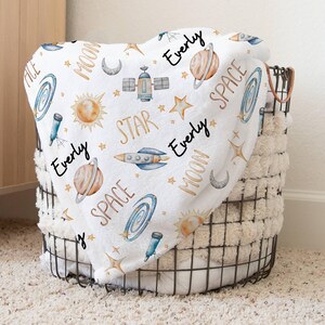 May include: A white baby blanket with a space theme. The blanket features a repeating pattern of planets, stars, rockets, and the words "Evely", "Moon", "Space", and "Star".