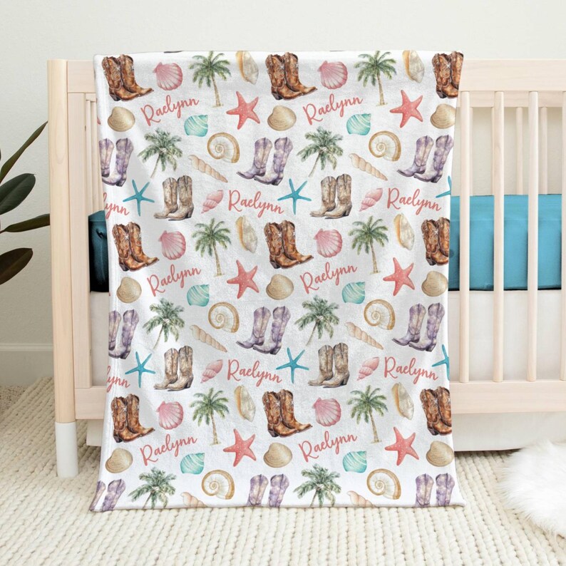 A white, beach themed baby blanket with a repeating pattern of pink cowgirl boots, palm trees, seashells, starfish, and the name 'Ralynn' draped off a crib. Blanket is colorful and designed in the coastal cowgirl aesthetic.