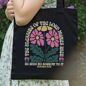 Puede incluir: Bolso tote negro con un diseño floral y el texto "THE BLESSING OF THE LORD MAKES RICH HE ADDS NO SORROW TO IT PROVERBS 10:22". El diseño incluye flores rosas y amarillas. El bolso lo sostiene una persona con un vestido floral.