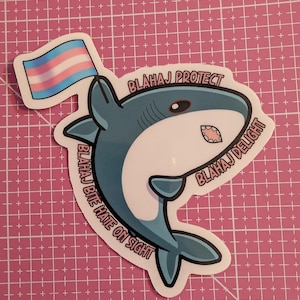 May include: A sticker featuring a cartoon shark holding a transgender pride flag. The shark is blue and white with the text "BLAHAJ PROTECT", "BLAHAJ DELIGHT", and "BLAHAJ BITE HATE ON SIGHT".