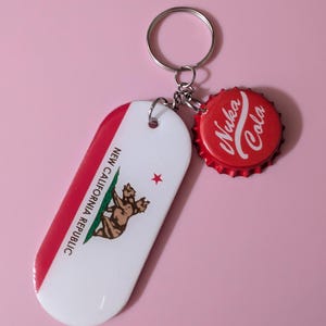 May include: A keychain featuring a metal ring attached to a red bottle cap with white Nuka Cola text and a white, red, and brown rectangular tag displaying the New California Republic flag with a two-headed bear and the text New California Republic.