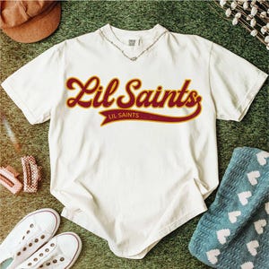 May include: Cream-colored t-shirt with the text "Lil Saints" in a maroon and gold script. The shirt is surrounded by accessories, including a brown hat, white sneakers, and a blue blanket with white hearts.