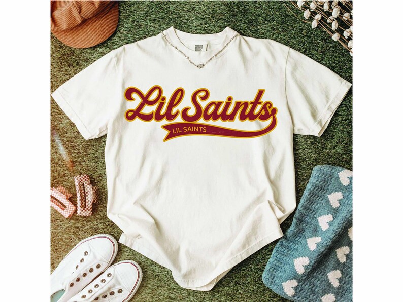 May include: Cream-colored t-shirt with the text "Lil Saints" in a maroon and gold script. The shirt is surrounded by accessories, including a brown hat, white sneakers, and a blue blanket with white hearts.