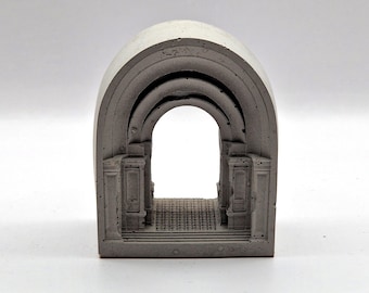 Puede incluir: Una escultura de arco de hormigón gris con un diseño arquitectónico detallado. La escultura presenta múltiples arcos, columnas y un suelo texturizado, creando una representación en miniatura de un edificio clásico. El arco abierto ofrece una vista clara a través de la estructura.