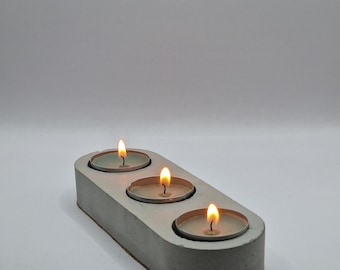 May include: A gray concrete candle holder with three lit tea lights. The holder is oval shaped and has a smooth, modern design.