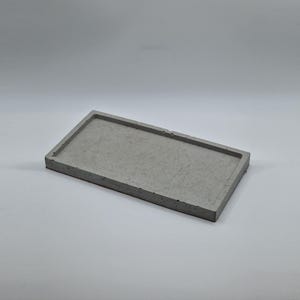 Puede incluir: Una bandeja rectangular de hormigón gris con un borde elevado. La bandeja es de un color sólido y neutro y tiene un diseño simple y minimalista. La bandeja es adecuada para guardar objetos pequeños.