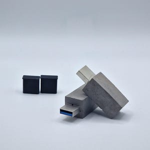 Peut inclure: Deux clés USB en béton avec un connecteur en métal argenté. Une clé est couchée sur le côté, tandis que l'autre est debout. Les clés sont sur une surface blanche.