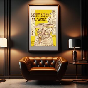 May include: Framed vintage poster with the text "Meet Me in St. Louis" in brown lettering. The poster features a yellow background with a trolley car illustration and a brown wooden frame. The poster is hanging above a brown leather armchair.
