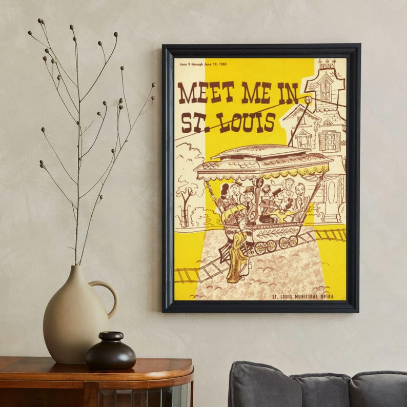 May include: Framed vintage poster with the text "Meet Me in St. Louis" in brown lettering on a yellow and tan background. The poster features a drawing of a trolley car with passengers and a woman in a long dress. The poster is in a black frame.