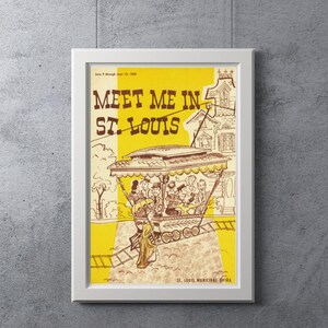 May include: A framed vintage poster with the text "Meet Me in St. Louis" in brown lettering on a yellow and cream background. The poster features an illustration of a trolley car with passengers and a building.