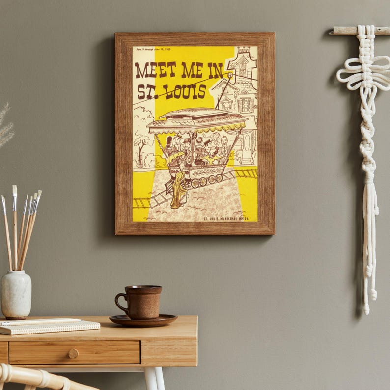 May include: A framed vintage poster with the text "Meet Me in St. Louis" in black letters on a yellow background. The poster features an illustration of a trolley car with passengers and a cityscape. The frame is made of wood.
