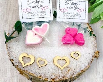 Pode incluir: Duas pequenas sacolas transparentes cheias de marshmallows em forma de coração rosa. Cada sacola tem um cartão branco com texto preto que diz "Herzklopfen in der Jüte" e "MARIA & FLORIAN 31. AUGUST 2024". Três recortes de madeira em forma de coração estão espalhados sobre uma superfície de madeira coberta de areia branca.