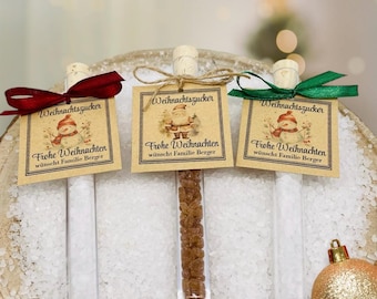 Puede incluir: Tres tubos de vidrio llenos de cristales de azúcar, cada uno con un corcho y una etiqueta decorativa. Las etiquetas dicen "Weihnachtszucker" y "Frohe Weihnachten" con una ilustración de muñeco de nieve o Papá Noel. Los tubos están atados con cintas rojas, verdes y marrones.