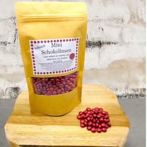 Peut inclure: Un sac de lentilles de chocolat rouge étiqueté "Mini Schokolinsen" avec le texte "Zum einfach so naschen oder dekorieren von Kuchen" et "100 Gramm". Le sac est posé sur une surface en bois avec un tas de lentilles de chocolat rouge devant lui.