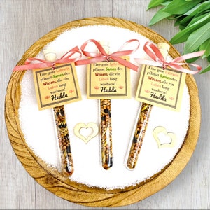May include: Three small glass tubes filled with seeds and tied with pink ribbon. Each tube has a small card attached with the text "Eine gute Lehrerin pflanzt Samen des Wissens, die ein Leben lang wachsen! Hedda".