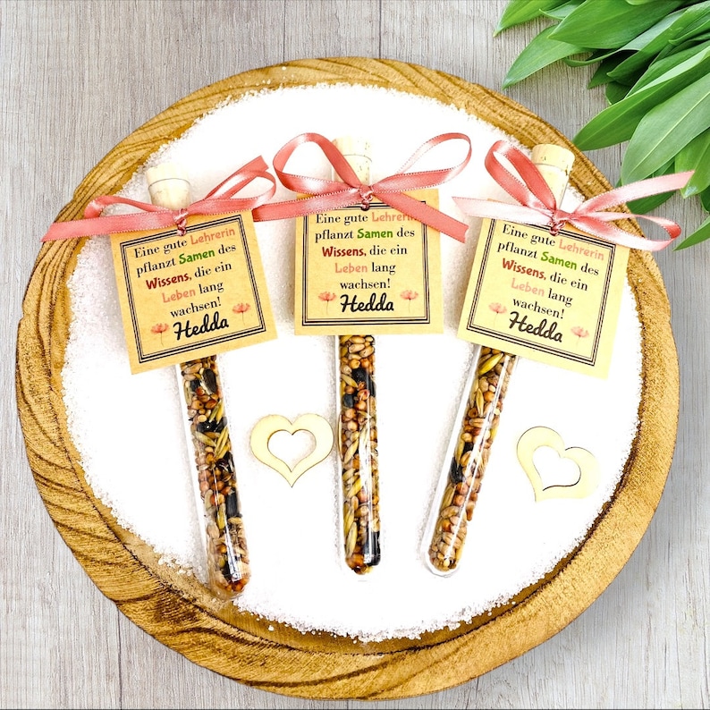 May include: Three small glass tubes filled with seeds and tied with pink ribbon. Each tube has a small card attached with the text "Eine gute Lehrerin pflanzt Samen des Wissens, die ein Leben lang wachsen! Hedda".