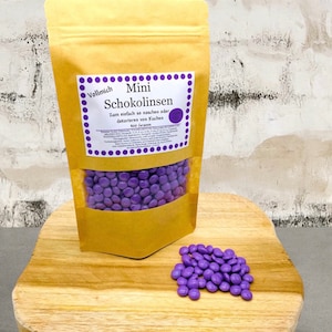 Könnte beinhalten: Ein brauner Papierbeutel gefüllt mit violetten Schokoladenlinsen. Der Beutel hat ein Etikett mit der Aufschrift "Vollmich Mini Schokolinsen Zum einfach so naschen oder dekorieren von Kuchen 400 Gramm".