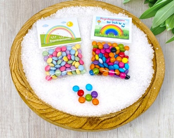 Op de afbeelding: Twee kleine zakjes met kleurrijke snoepjes met regenboogthema met de tekst "Regenbogen samen", wat "Regenboog samen" betekent. De zakjes liggen op een houten kom gevuld met wit zand. Er liggen een paar losse snoepjes op het zand.