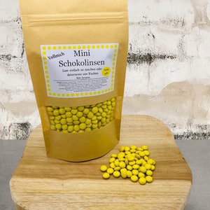 Könnte beinhalten: Ein brauner Papierbeutel gefüllt mit gelben Schokoladenlinsen. Der Beutel hat ein Etikett mit der Aufschrift "Vollmich Mini Schokolinsen Zum einfach so naschen oder dekorieren von Kuchen 400 Gramm". Auf einer Holzoberfläche liegen auch gelbe Schokoladenlinsen verstreut.
