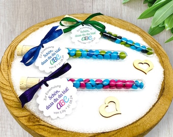 May include: Three glass test tubes filled with colorful candy, each with a white tag attached. The tags have a scalloped edge and say "Zur Einschulung" which translates to "For the start of school" in English. The tags also have the text "Schön, dass du da bist!" which translates to "It's great that you're here!" in English. Each tag has a different name and date.