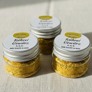 Puede incluir: Tres pequeños frascos de vidrio llenos de una mezcla de especias amarilla. Cada frasco tiene una tapa plateada y una etiqueta blanca con texto en alemán. La etiqueta dice "Rührei Gewürz" y "Der kleine für Dich".
