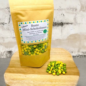 Könnte beinhalten: Eine Tüte mit gelben und grünen Schokoladenlinsen namens "Bunte Mini-Schokolinsen". Die Tüte liegt auf einer Holzoberfläche mit einem Haufen Schokoladenlinsen davor. Auf der Tüte steht "Zum einfach so naschen oder dekorieren von Kuchen" und "400 Gramm".
