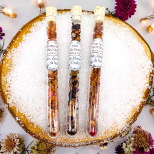 Op de afbeelding: Drie glazen buisjes gevuld met gedroogd fruit en thee, elk afgesloten met een kurk. De buisjes zijn gelabeld met Duitse tekst en liggen op een houten schijf bedekt met witte kristallen. Bloemenaccenten en feeërieke lichtjes omringen de buisjes.