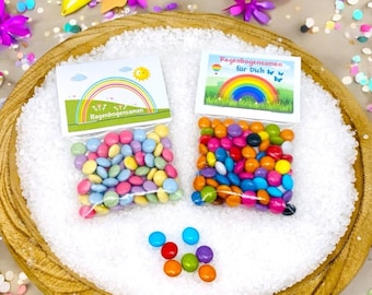 Op de afbeelding: Twee kleine zakjes met kleurrijke snoepjes met regenboogthema met de tekst "Regenbogensamen" en "Regenbogensamen fur Dich". De zakjes liggen op een houten bord met witte sprinkles.