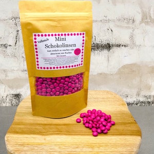 Könnte beinhalten: Eine Tüte mit rosa Mini-Schokoladenlinsen mit der Aufschrift "Vollmich Mini Schokolinsen Zum einfach so naschen oder dekorieren von Kuchen 400 Gramm". Die Tüte liegt auf einer Holzoberfläche mit einem kleinen Haufen rosa Schokoladenlinsen davor.