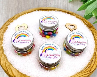 Op de afbeelding: Drie kleine glazen potjes gevuld met kleurrijke snoepjes, elk met een zilveren deksel en een label met eenhoorn-thema met de tekst "Schen, dass du da warst! 6. Geburtstag von Lilly". De potjes staan op een houten dienblad gevuld met wit zand.