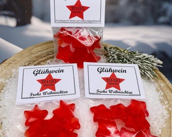 Puede incluir: Bolsas transparentes llenas de caramelos de goma rojos en forma de estrella. Cada bolsa está etiquetada con "Glühwein TO GO" y "Frohe Weihnachten". Los caramelos están sobre una cama de cristales blancos.