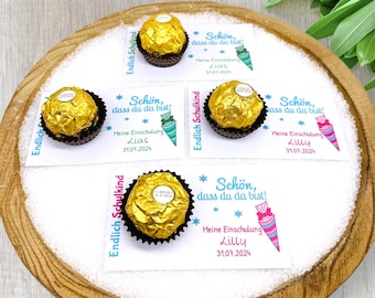 May include: Four white gift tags with blue and pink stars and a blue and pink school cone design. Each tag has the text "Endlich Schulkind" and "Schön, dass du da bist!" with a name and date. The tags are attached to chocolate balls wrapped in gold foil.