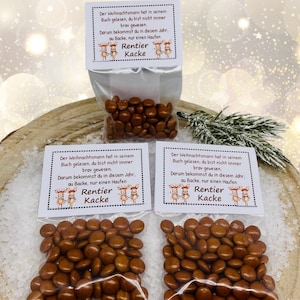 Può includere: Piccoli sacchetti trasparenti pieni di caramelle rotonde marroni. Ogni sacchetto ha un'etichetta con testo in tedesco e una grafica di renna. I sacchetti sono disposti su una superficie di legno con uno sfondo innevato.