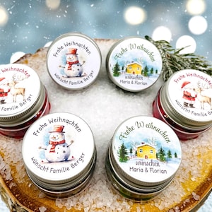 May include: Several small, round metal tins with Christmas-themed lids are displayed. The lids are decorated with illustrations of snowmen, Santa Claus with a reindeer, and a winter scene with a house. The text "Frohe Weihnachten" is printed on the lids.