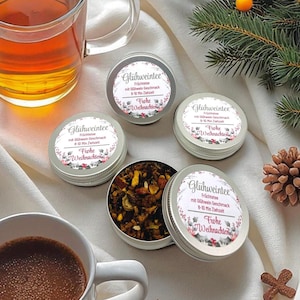 Può includere: Una composizione festosa con piccole scatole d'argento di Glühweintee, alcune aperte per rivelare le foglie di tè. Sono presenti due tazze di tè e una tazza di caffè, insieme ad aghi di pino, pigne e una stella di legno. Le scatole hanno etichette con testo.